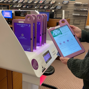 A person holds a purple Hublet tablet in front of a self-service kiosk at Clarkston Library. The station holds five additional tablets in protective purple cases.